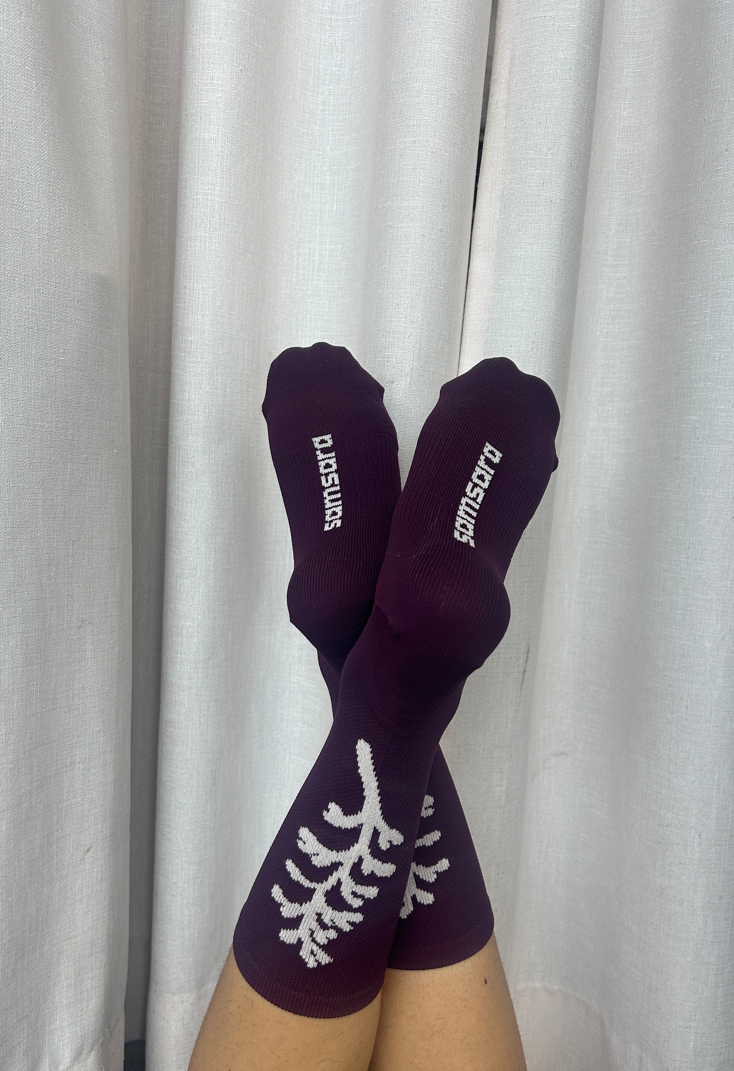 A pair of dark purple crew socks with white knit tree branch patterns and the brand name 'pomsutra' printed in white on the toes, shown on feet against a white curtain background.