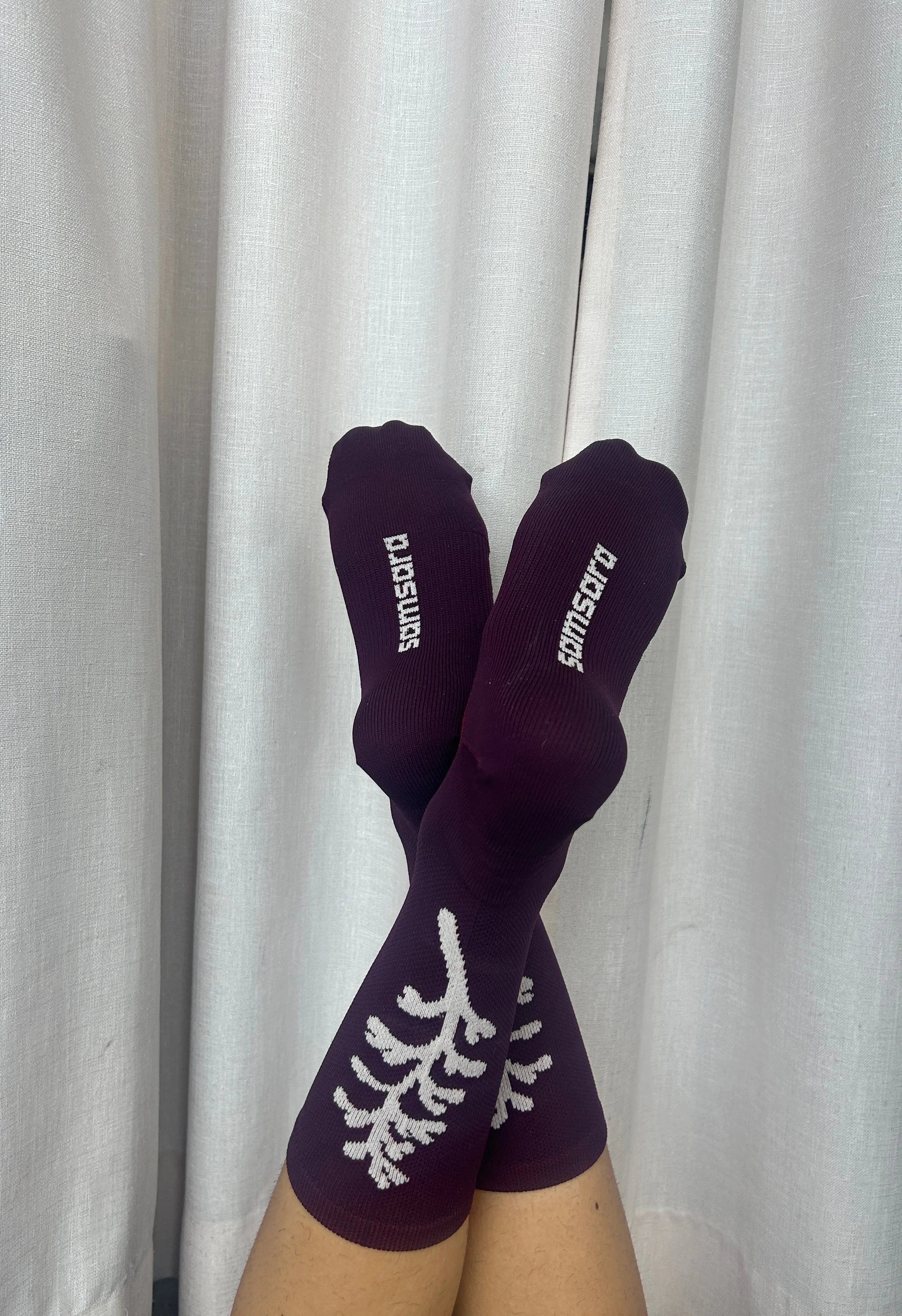 A pair of dark purple crew socks with white knit tree branch patterns and the brand name 'pomsutra' printed in white on the toes, shown on feet against a white curtain background.
