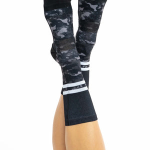 Top view of women’s crew cycling socks with black and grey camo on the bottom and black top with white stripes on a women’s feet.